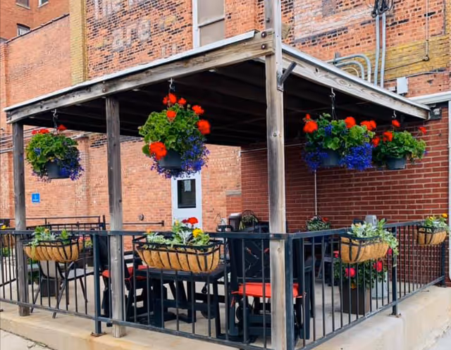 Outdoor patio area with a wooden pergola roof, black metal railing, and several hanging flower baskets with red and purple flowers. There are black chairs with red cushions and a table underneath the pergola, set against a brick building wall.