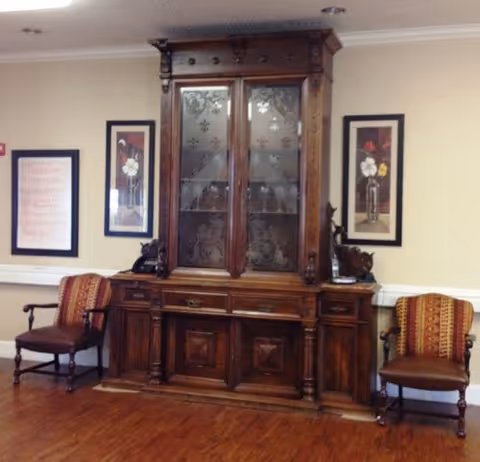 An interior room featuring a large, ornate wooden cabinet with glass doors etched with floral designs. On either side of the cabinet are two wooden armchairs with patterned upholstery in red and gold tones. The walls are light-colored and decorated with three framed pictures, two of which depict flowers in vases. The floor is wooden.
