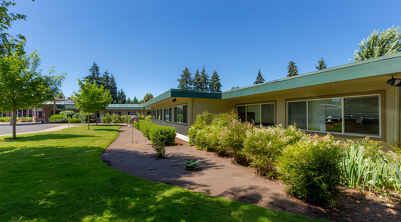 Exterior view of a single-story building with large windows, surrounded by green grass, bushes, and trees under a clear blue sky.
