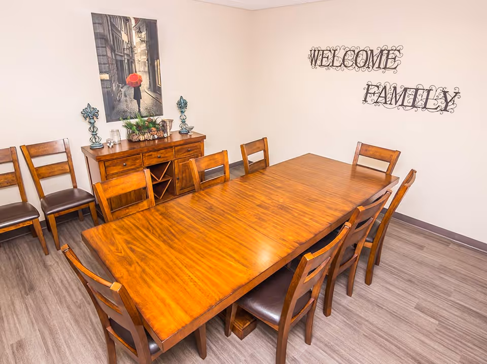 A dining room with a large wooden table surrounded by eight wooden chairs with dark cushions. Against the wall is a wooden sideboard with decorative items including two blue candle holders, a glass jar, and a centerpiece with greenery and pinecones. Above the sideboard hangs a framed picture of a person holding a red umbrella walking down a narrow street. On the adjacent wall, decorative metal letters spell out 'WELCOME FAMILY'. The room has light-colored walls and wood-look flooring.