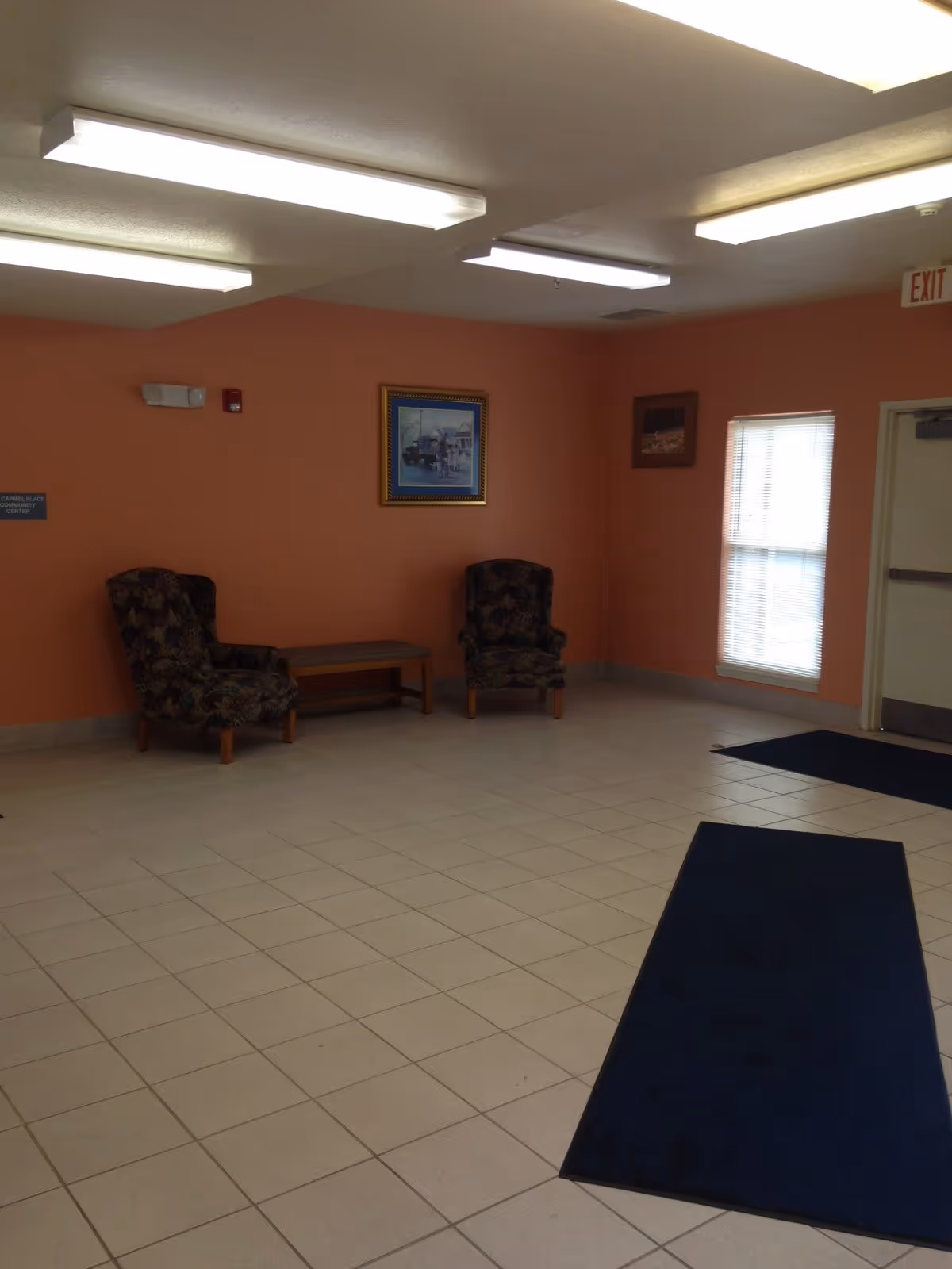 A waiting area with two patterned armchairs and a wooden bench against a peach-colored wall. There are two framed pictures hanging on the wall, a window with blinds, and an exit door. The floor is tiled with light-colored tiles and has two dark blue rugs. The ceiling has several fluorescent lights.