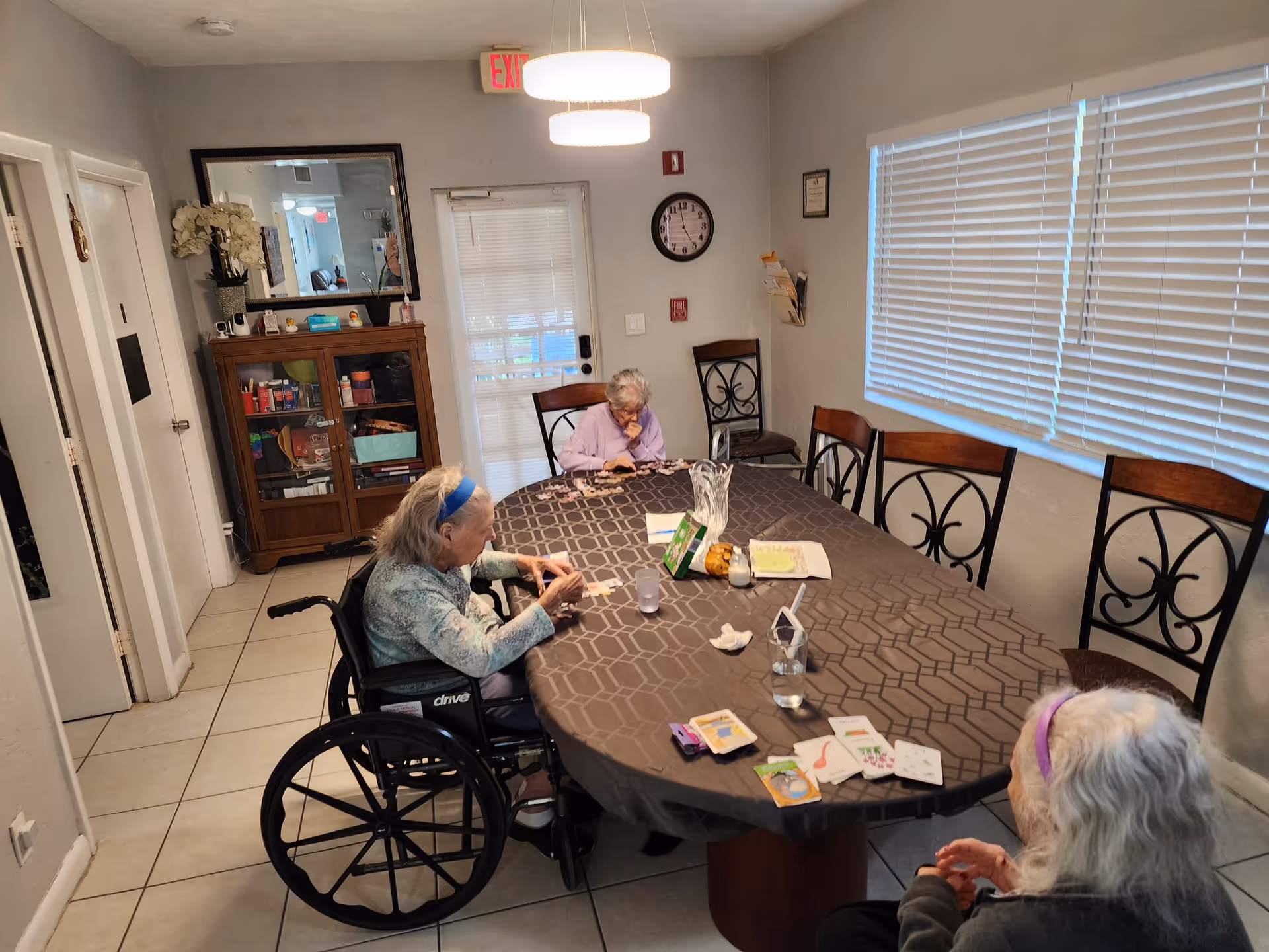 Three elderly women sitting around a large oval table in a well-lit room with a window covered by blinds. One woman is in a wheelchair, and they appear to be engaged in an activity involving cards or small items on the table. The room has tiled floors, a wooden cabinet with glass doors, a wall clock, and a door with a window covered by blinds.
