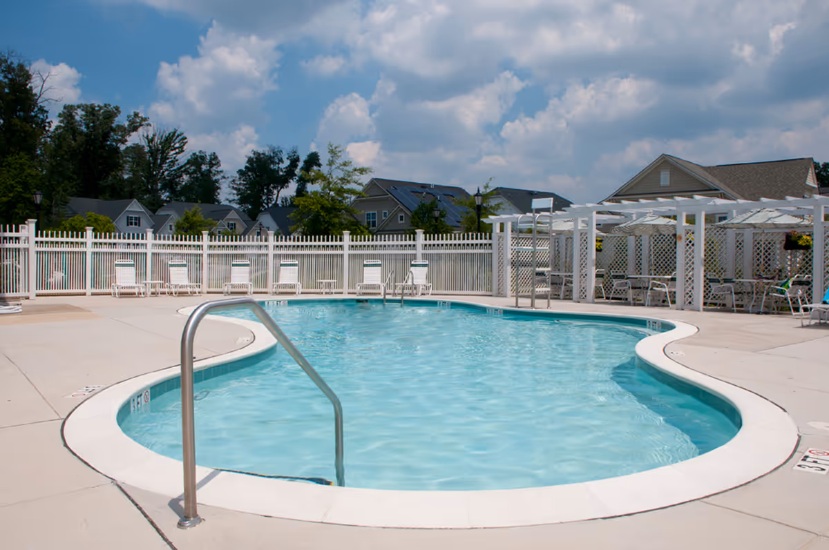 Outdoor swimming pool with a white fence, lounge chairs, and a pergola-covered seating area at a residential facility under a partly cloudy sky.