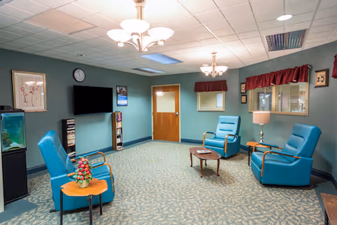 A cozy common area with teal armchairs arranged around small wooden tables, a wall-mounted flat-screen TV, a fish tank, and framed artwork on the walls. The room has a patterned carpet, ceiling lights, and a door leading to another room.