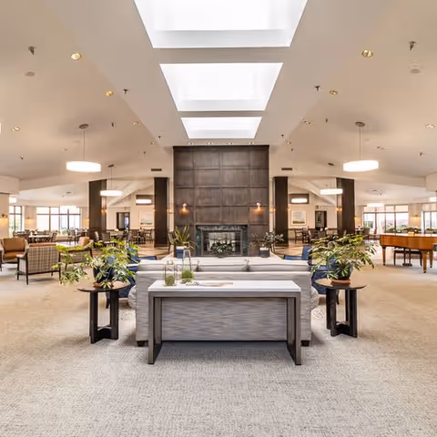 Spacious and well-lit senior living facility common area with a large central fireplace, comfortable seating including sofas and armchairs, side tables with plants, and skylights overhead providing natural light. The room has a neutral color palette with beige carpeting and modern light fixtures.