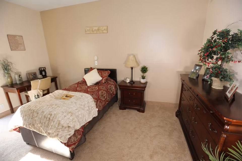 A neatly decorated single bedroom with a bed, nightstand and lamp, a dresser topped with plants and photos, and a small desk.