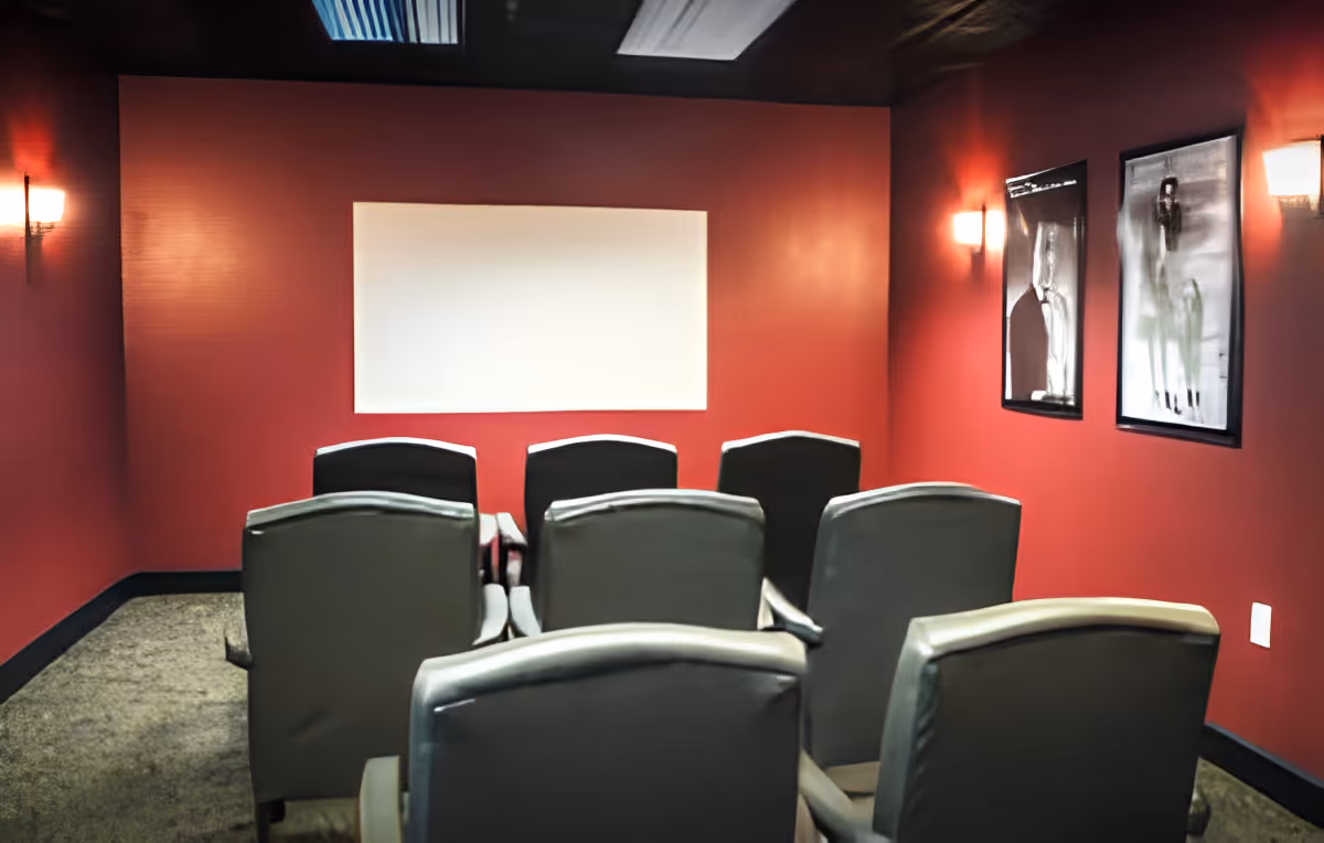 Small theater room with red walls, eight gray cushioned chairs arranged in two rows facing a blank white screen, wall sconces providing soft lighting, and two framed abstract artworks on the right wall.