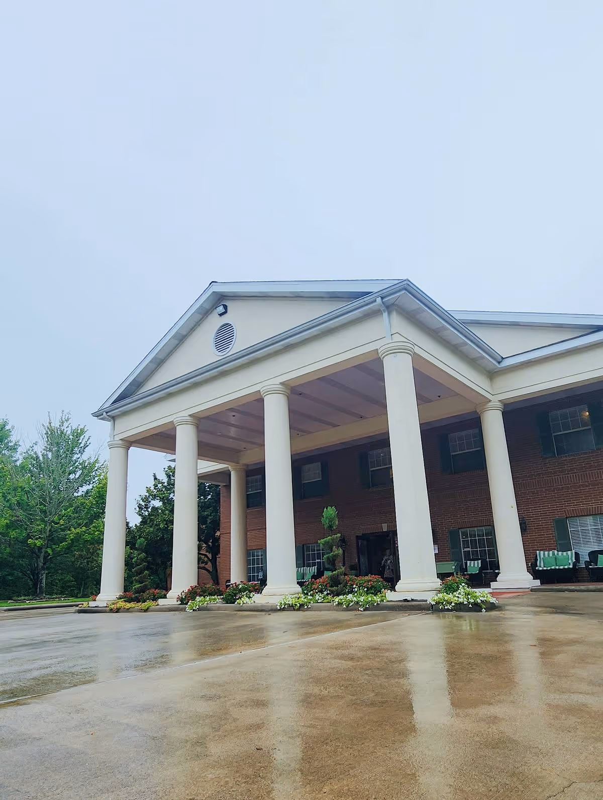 Front exterior view of a building with large white columns supporting a covered entrance. The building has a brick facade with multiple windows and green shutters. There are plants and flowers arranged at the base of the columns, and the ground appears wet from rain. Trees are visible in the background under an overcast sky.