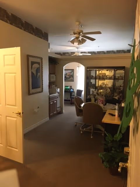 Interior view of a senior living facility room with an open door on the left, a ceiling fan, a small kitchenette area, a glass display cabinet, chairs around a table, and some framed artwork on the walls.