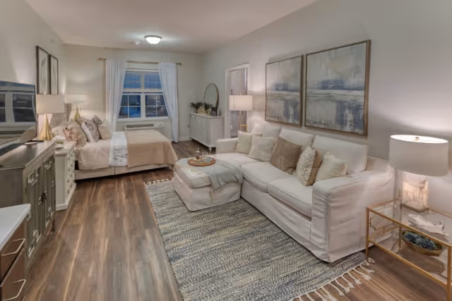 A cozy and well-lit interior room featuring a white sofa with multiple cushions, a matching ottoman with a tray, a large area rug, two abstract paintings on the wall, a side table with a lamp, and a bed with pillows and a beige throw blanket near a window with white curtains.