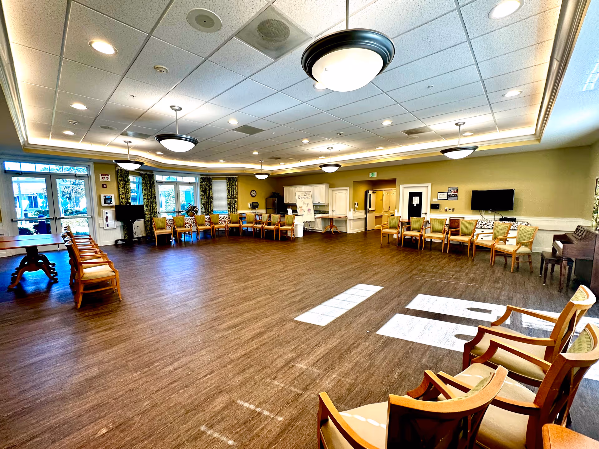 Spacious senior living communal activity room with chairs arranged around the perimeter, large windows, and overhead lighting.