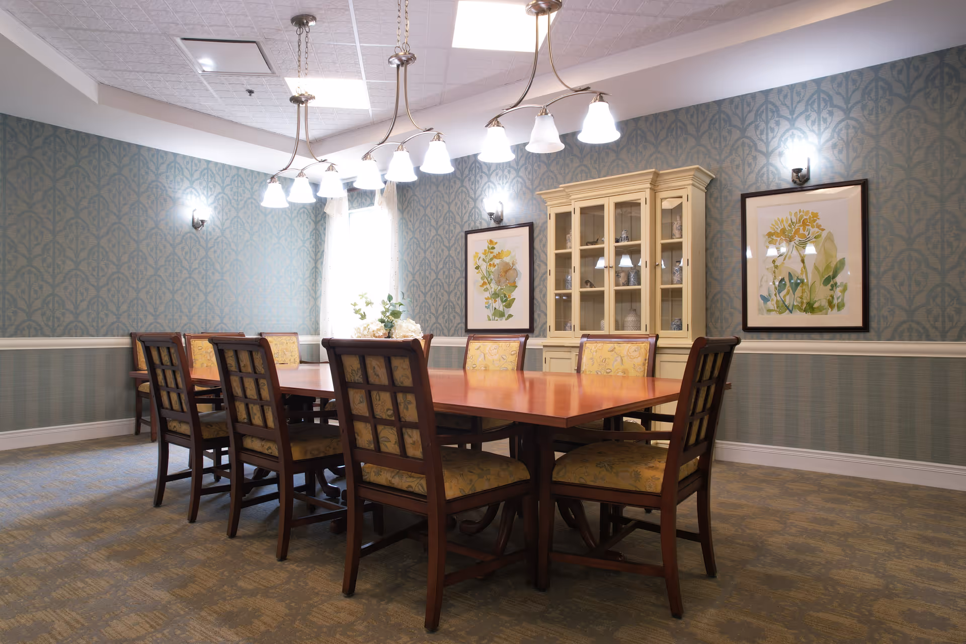 A formal dining room with a long wooden table surrounded by eight cushioned chairs. The walls are decorated with patterned wallpaper and two framed botanical prints. A cream-colored china cabinet stands against the wall, and a chandelier with multiple lights hangs above the table. The room has a carpeted floor and soft lighting.