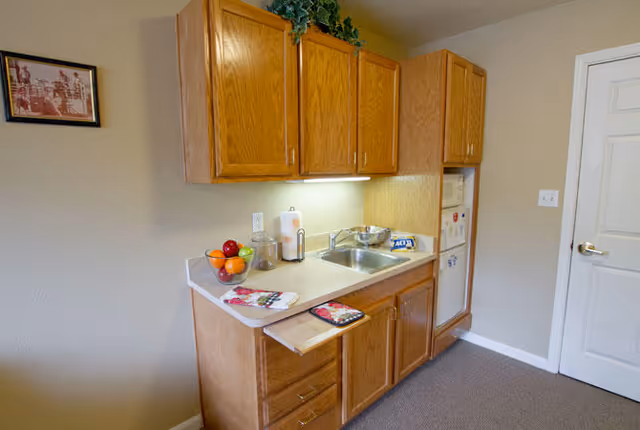A small kitchen area with wooden cabinets, a countertop with a sink, a bowl of fruit, a paper towel holder, and a microwave. There is a white door to the right and a framed picture on the wall to the left.