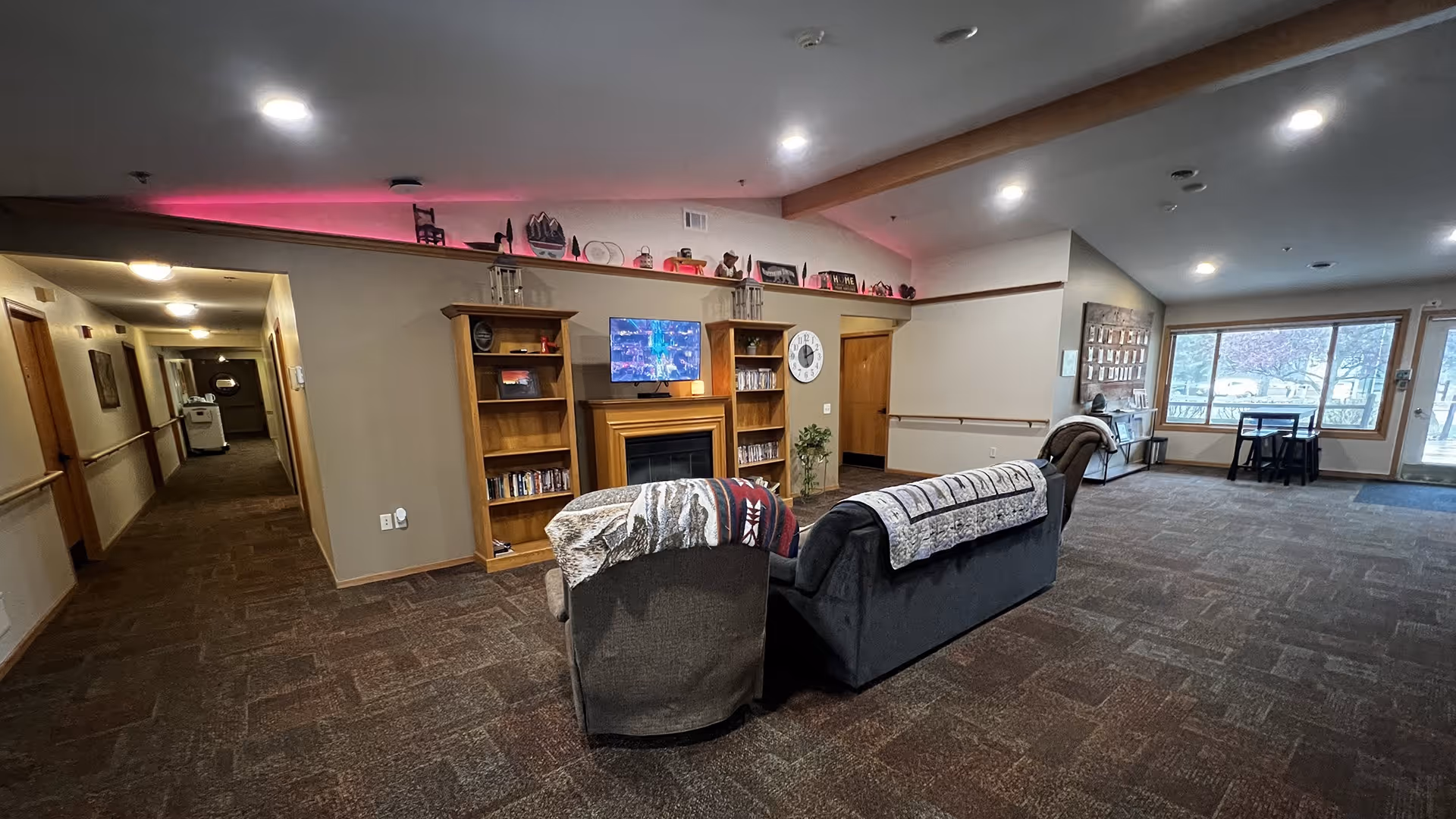 Spacious assisted-living common room with sofas facing a fireplace and TV, bookshelves, a hallway to the left, and large windows to the right.
