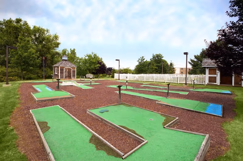 Outdoor miniature golf course with several green putting areas bordered by wood, surrounded by brown mulch and grass. There are small buildings, trees, and a white picket fence in the background under a partly cloudy sky.
