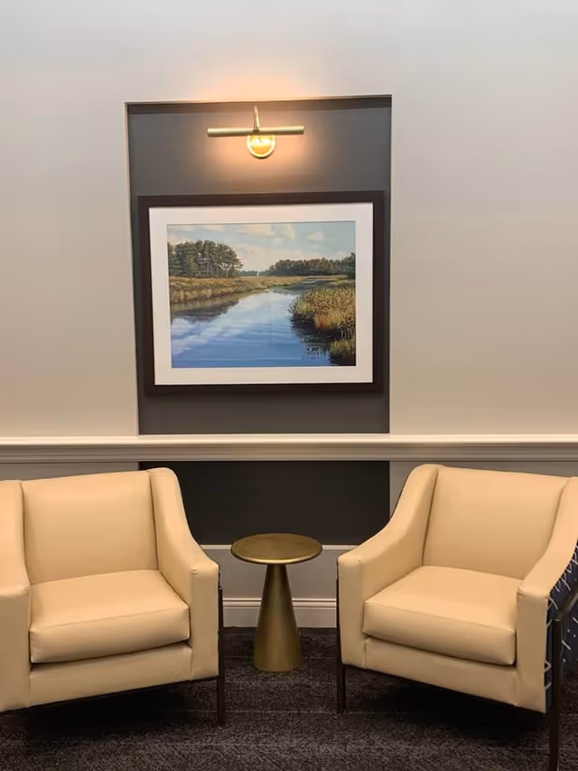 Two beige armchairs flanking a small round brass table beneath a framed landscape painting and wall sconce in a seating area.