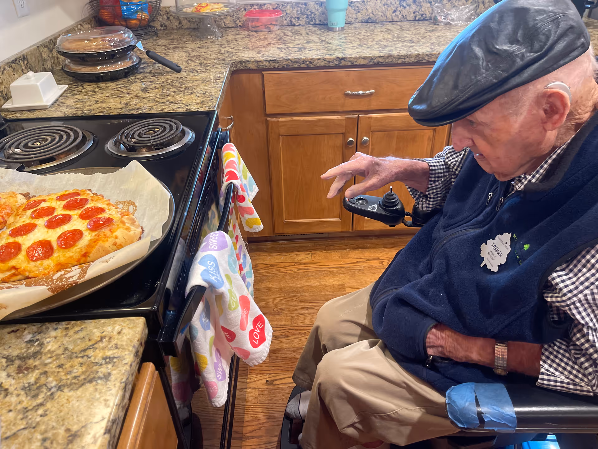 An elderly man in a wheelchair wearing a black cap and navy vest is sitting in a kitchen next to a stove with a freshly baked pepperoni pizza on a baking tray. The kitchen has wooden cabinets and granite countertops, with colorful towels hanging on the oven handle.