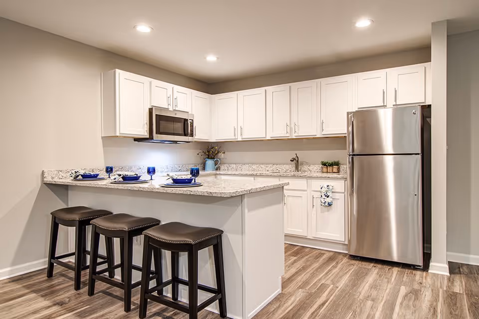 Modern kitchen with white cabinets, granite countertops, stainless steel refrigerator and microwave, three black bar stools at the kitchen island set with blue plates and glasses, and wood-style flooring.