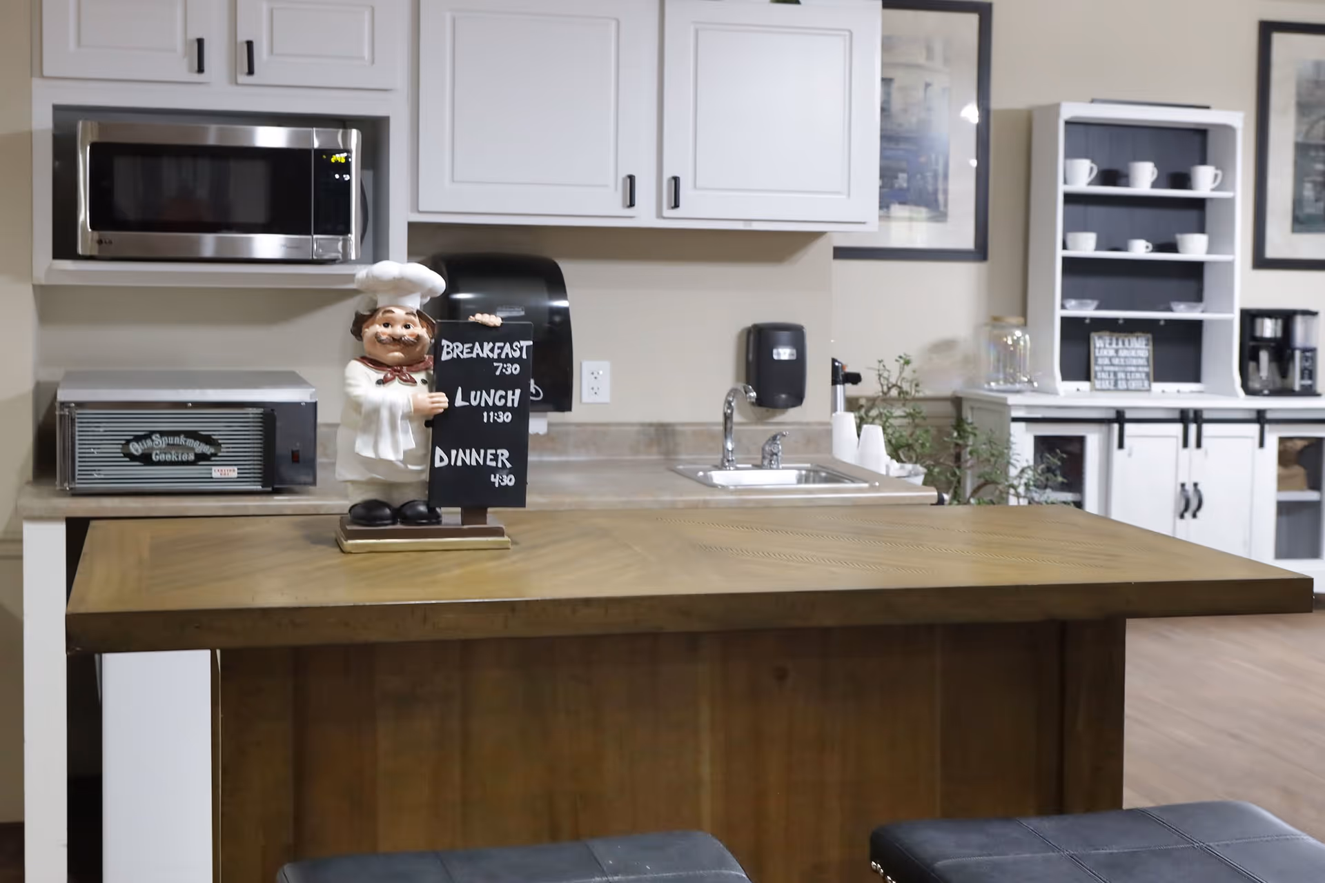 A kitchen area with a wooden counter in the foreground, a microwave and toaster oven on the countertop, white cabinets above, a sink with a soap dispenser, and a small chef figurine holding a chalkboard sign listing meal times. In the background, there is a white hutch with cups and a coffee maker.