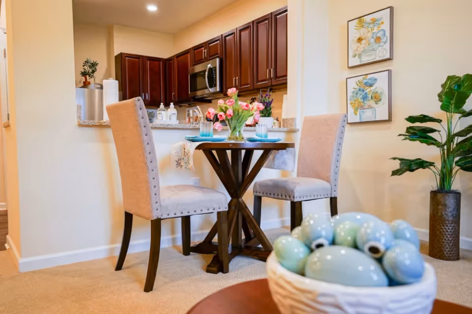 A cozy dining area with a small round wooden table set for two with plates, glasses, and a vase of pink tulips. Two beige upholstered chairs with nailhead trim are positioned around the table. Behind the dining area is a kitchen with dark wooden cabinets, a microwave, and a granite countertop. The wall features two framed floral paintings, and a tall green plant is placed in a decorative pot on the right side. In the foreground, there is a blurred basket filled with decorative blue and green ceramic eggs.