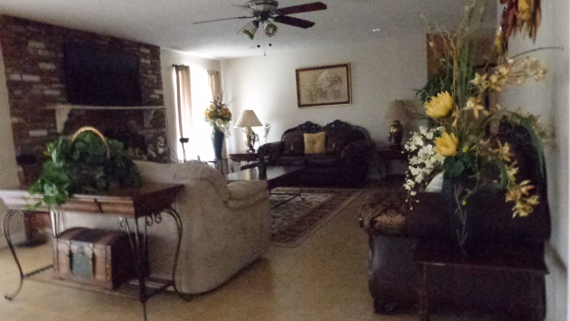 A cozy living room with a beige sofa, dark leather armchairs, a wooden coffee table, and a patterned rug. There is a brick fireplace with a mounted TV above it, a ceiling fan with lights, and several floral arrangements placed around the room. The walls are light-colored, and there are windows with curtains letting in natural light.