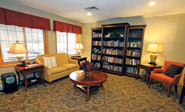 A cozy living room area with a beige loveseat, a red patterned armchair, two wooden side tables with lamps, a round wooden coffee table with a flower arrangement, and a large bookshelf filled with books against the wall. The room has carpeted flooring and two windows with red valances letting in natural light.