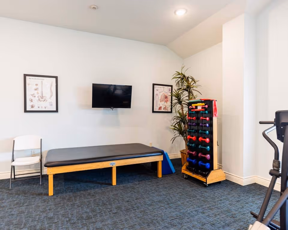 A small exercise room with a padded wooden therapy table, a white folding chair, a wall-mounted flat screen TV, anatomical posters on the wall, a rack holding colorful dumbbells, a potted plant, and part of an exercise machine visible on the right.