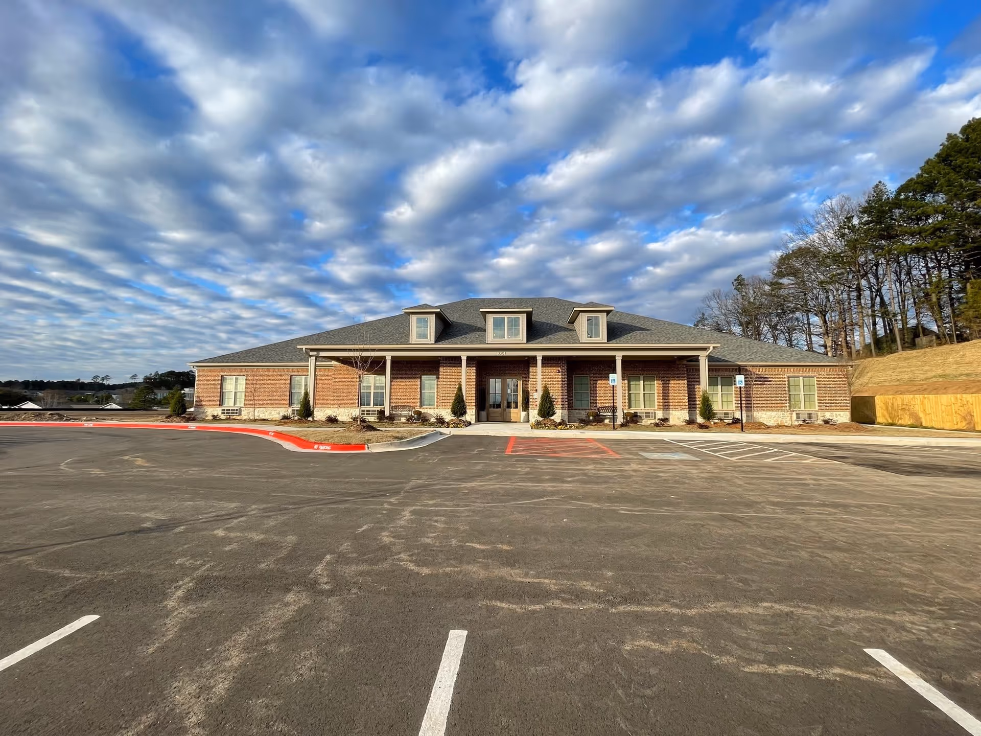 Front exterior view of a single-story brick building with a gray shingled roof and three dormer windows. The building has a covered entrance supported by columns and is surrounded by a paved parking lot with marked parking spaces and handicap signs. Trees and a partly cloudy sky are visible in the background.