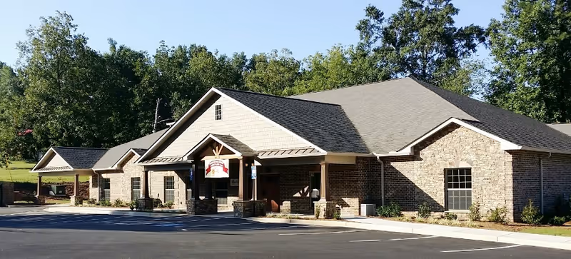Exterior view of a single-story brick building with a covered entrance and a sign hanging above it, surrounded by trees and a parking lot in front.