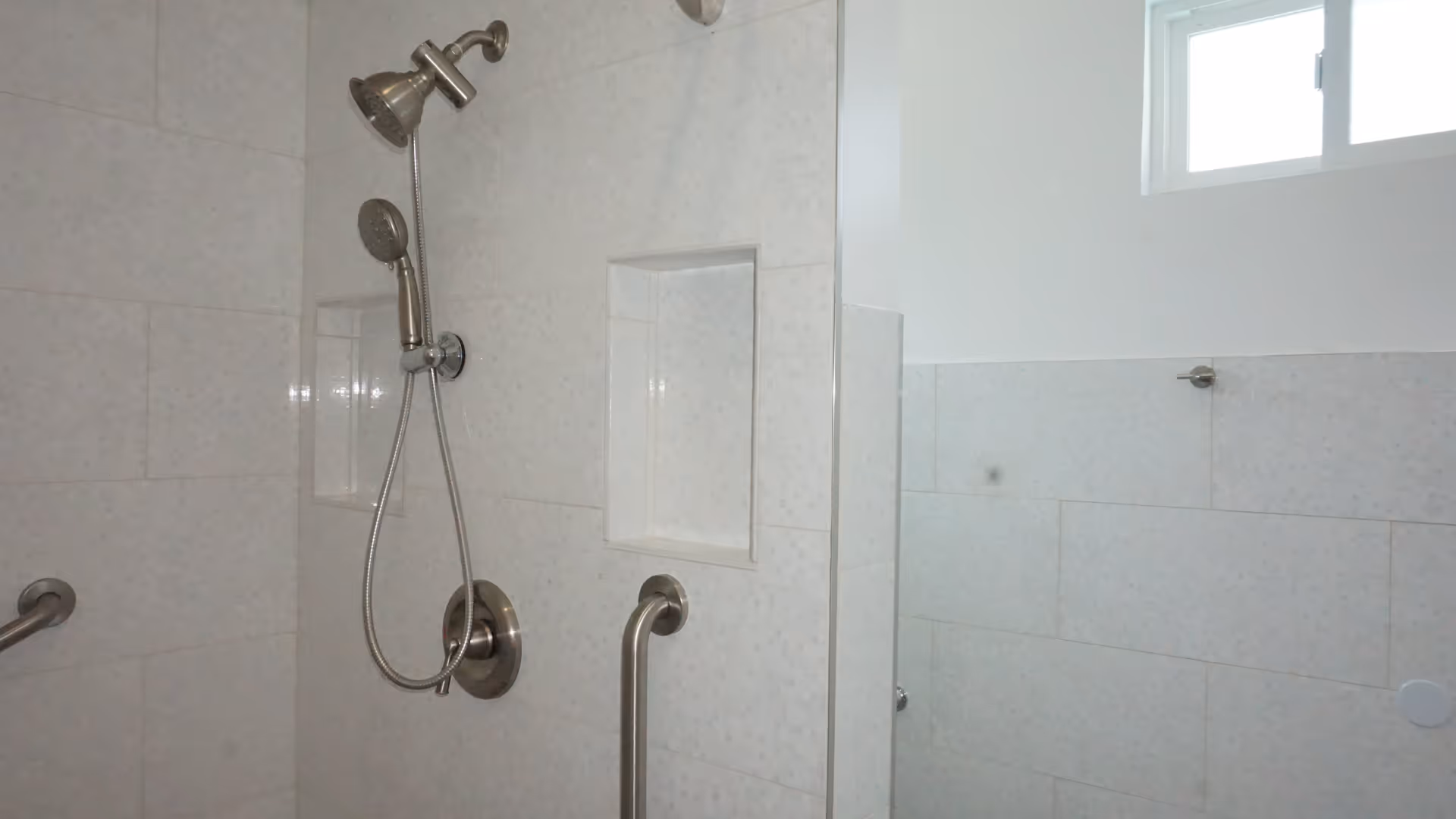 A tiled shower area with a handheld showerhead and a fixed showerhead mounted on the wall. There are two built-in wall niches for toiletries and a grab bar for safety. A small window is visible near the ceiling, allowing natural light into the space.