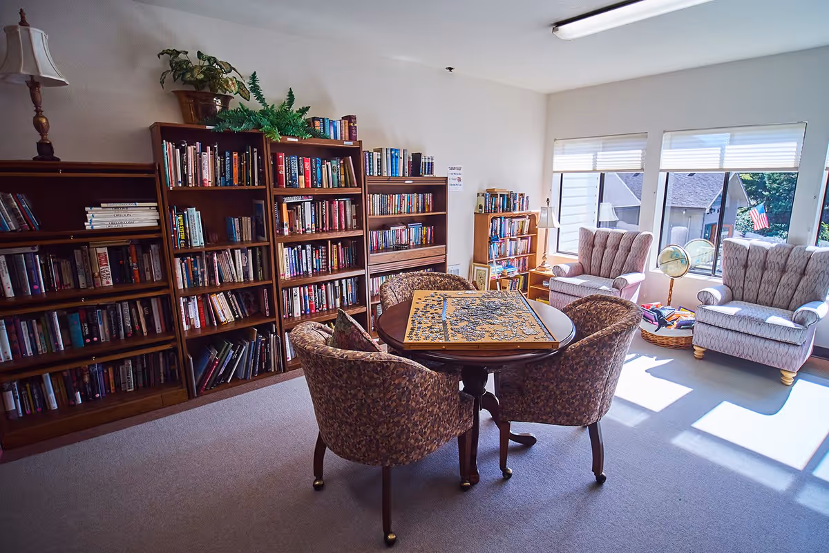 A cozy reading and puzzle area in a senior living facility with bookshelves filled with books, a round table with an incomplete jigsaw puzzle, four cushioned chairs around the table, two armchairs near large windows letting in natural light, a globe, and a basket with books on the floor.