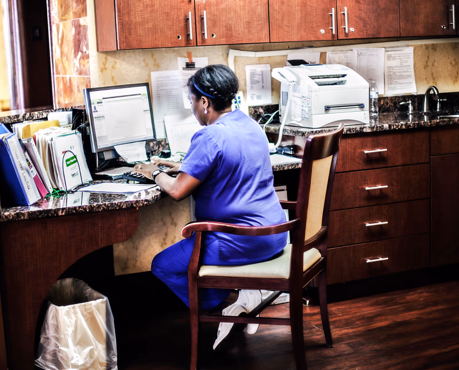 A caregiver in purple scrubs sits at a desk using a computer at a nursing station.