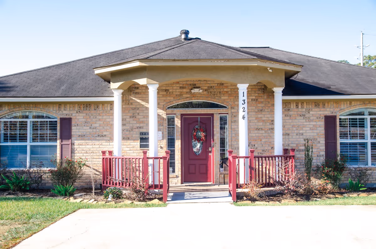 Front exterior view of a single-story brick building with a dark shingled roof, white columns supporting a small porch with red railings, and a maroon front door decorated with a wreath. The building number 1324 is displayed on one of the columns. There are windows with white shutters on either side of the door and some landscaping with bushes and plants in front.