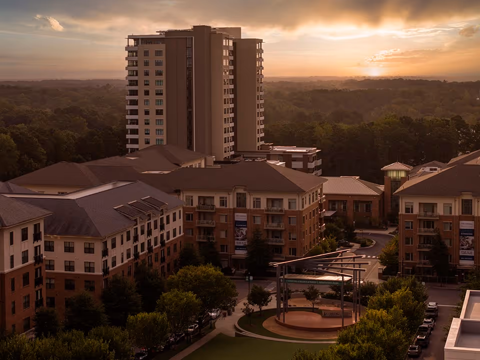Aerial view of The Cardinal at North Hills senior living facility at sunset, showing multiple multi-story residential buildings surrounded by trees and a small outdoor amphitheater with a circular stage and metal canopy structure.