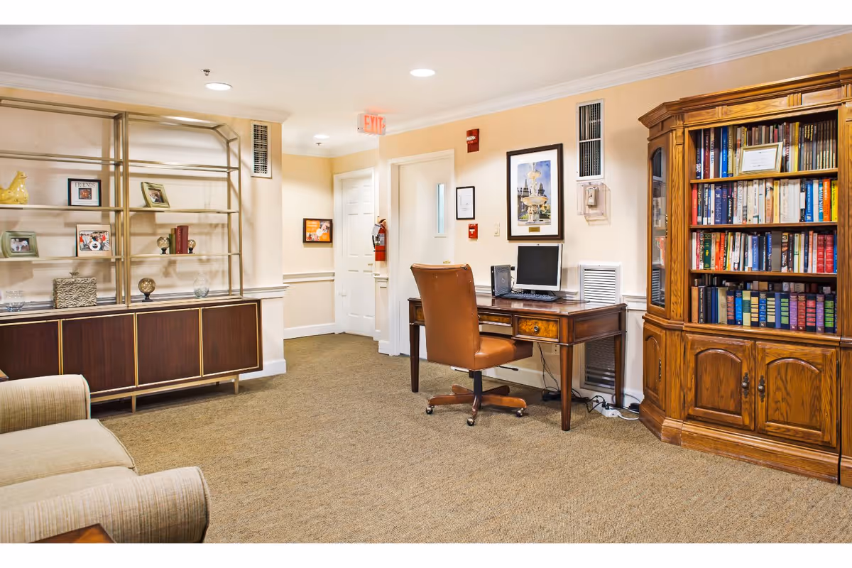 Interior common room with a wooden desk and computer, leather desk chair, large bookcase, display cabinet and upholstered seating.
