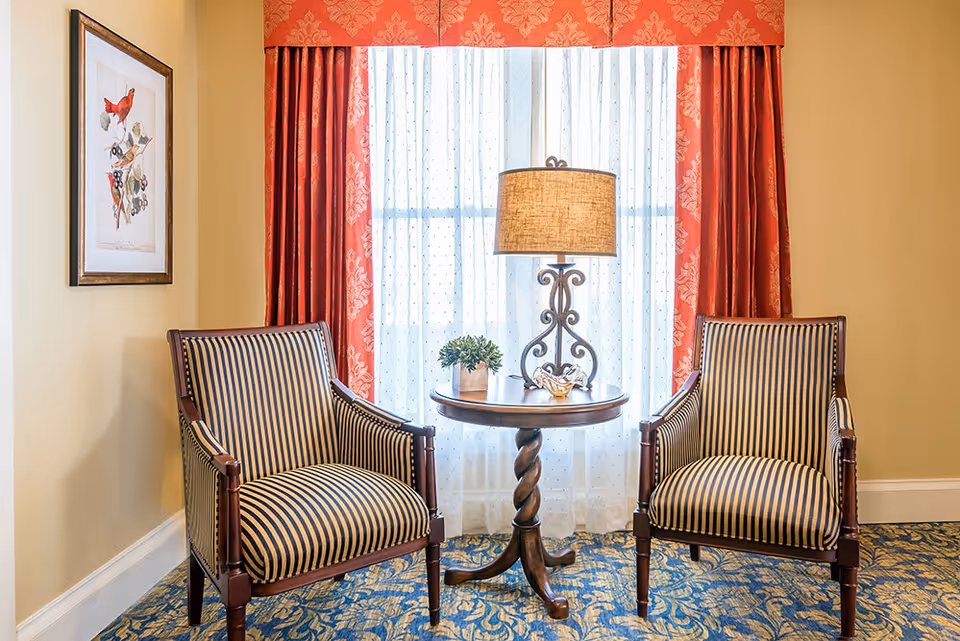 Two striped armchairs with wooden frames are positioned on either side of a small round wooden table with a twisted pedestal base. On the table, there is a decorative lamp with a beige shade, a small potted plant, and a glass bowl. Behind the table is a window with sheer white curtains and red patterned drapes. A framed picture of birds hangs on the left wall. The room has beige walls and a blue patterned carpet.