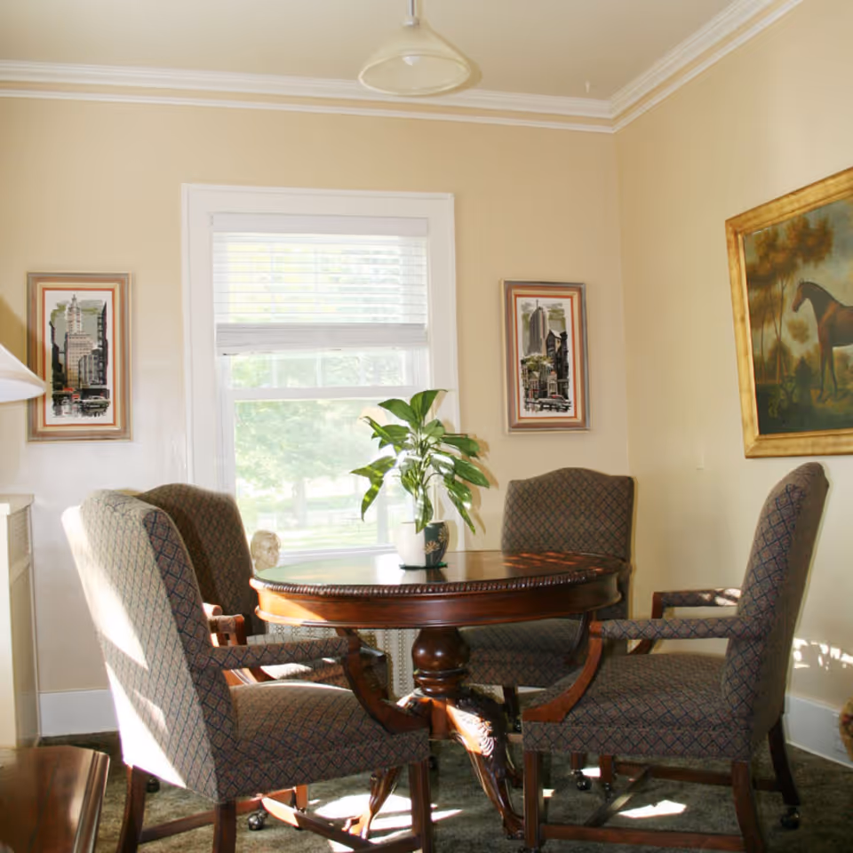 A cozy room with a round wooden table surrounded by four upholstered armchairs. A potted plant sits on the table. The room has beige walls with three framed artworks, including a painting of a horse and two cityscape prints. A window with white blinds lets in natural light.