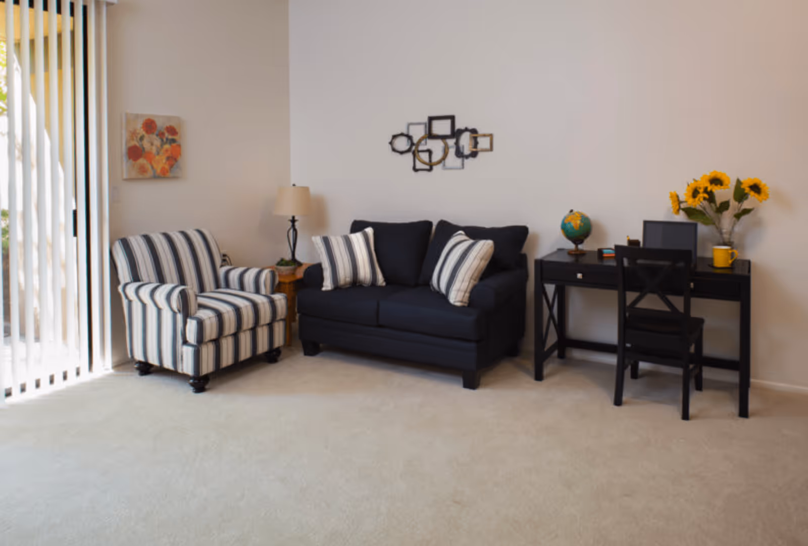 A cozy living room area with a striped armchair, a dark blue loveseat with two striped pillows, a small wooden side table with a lamp, and a black desk with a chair. On the desk are a globe, a laptop, a yellow mug, and a vase with sunflowers. The room has light-colored carpet and walls, with vertical blinds covering a sliding glass door on the left side.