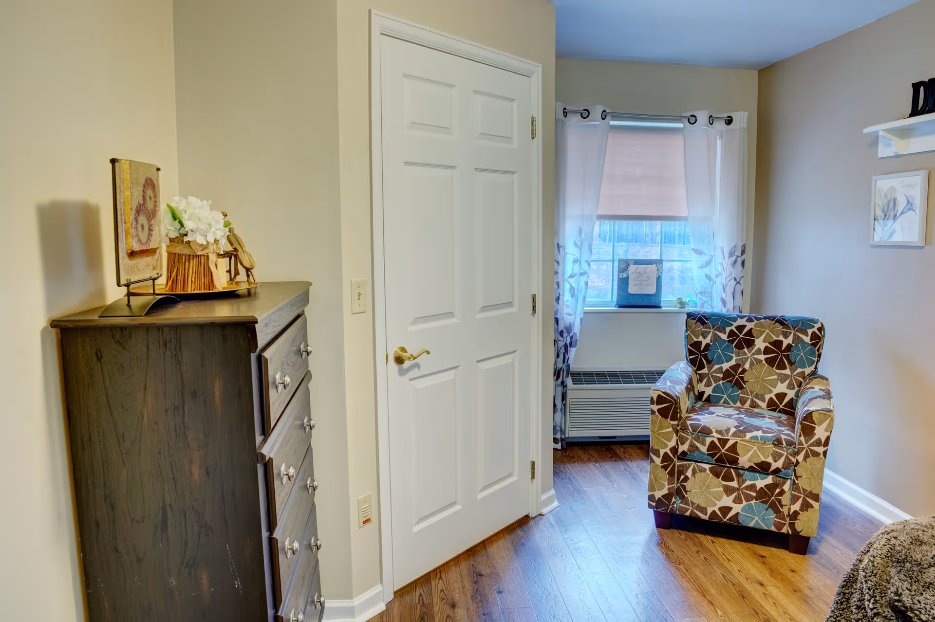A cozy corner of a room featuring a patterned armchair with floral designs, a window with sheer white curtains and a beige roller shade, a dark wooden chest of drawers with decorative items on top, and a closed white door. The room has light beige walls and wooden flooring.