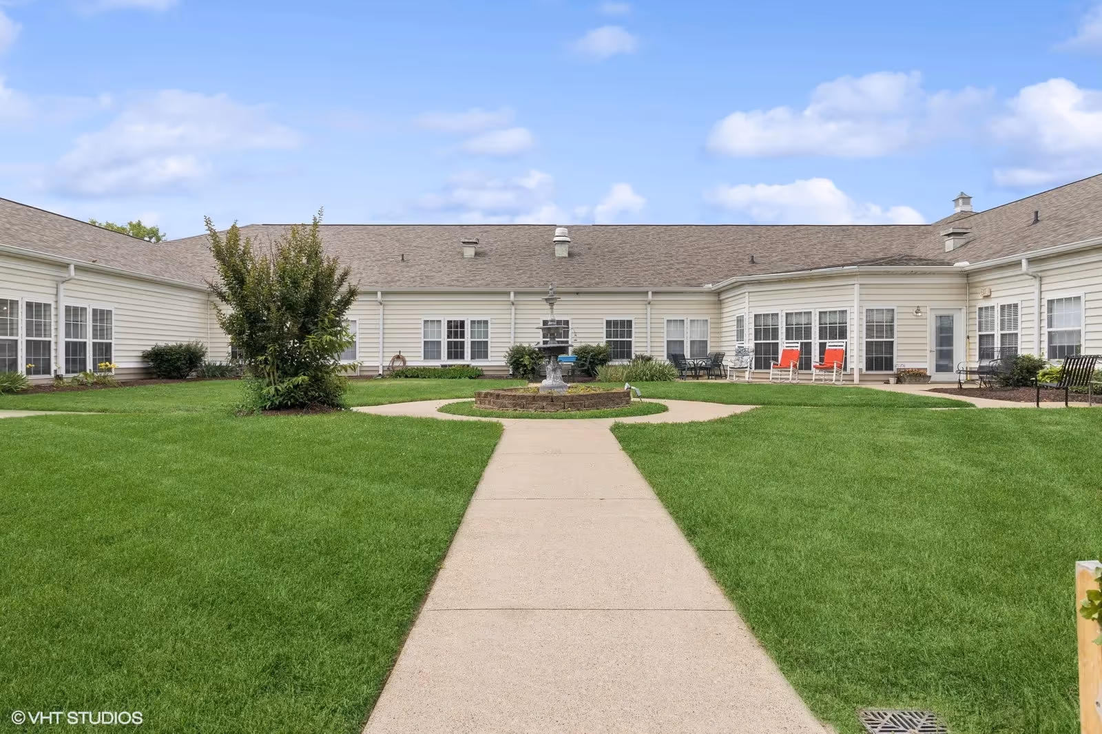 A courtyard area of a senior living facility with a concrete walkway leading to a central fountain. The building surrounds the courtyard with multiple windows and doors. There are green lawns, some bushes, and outdoor seating including chairs and benches. The sky is partly cloudy.