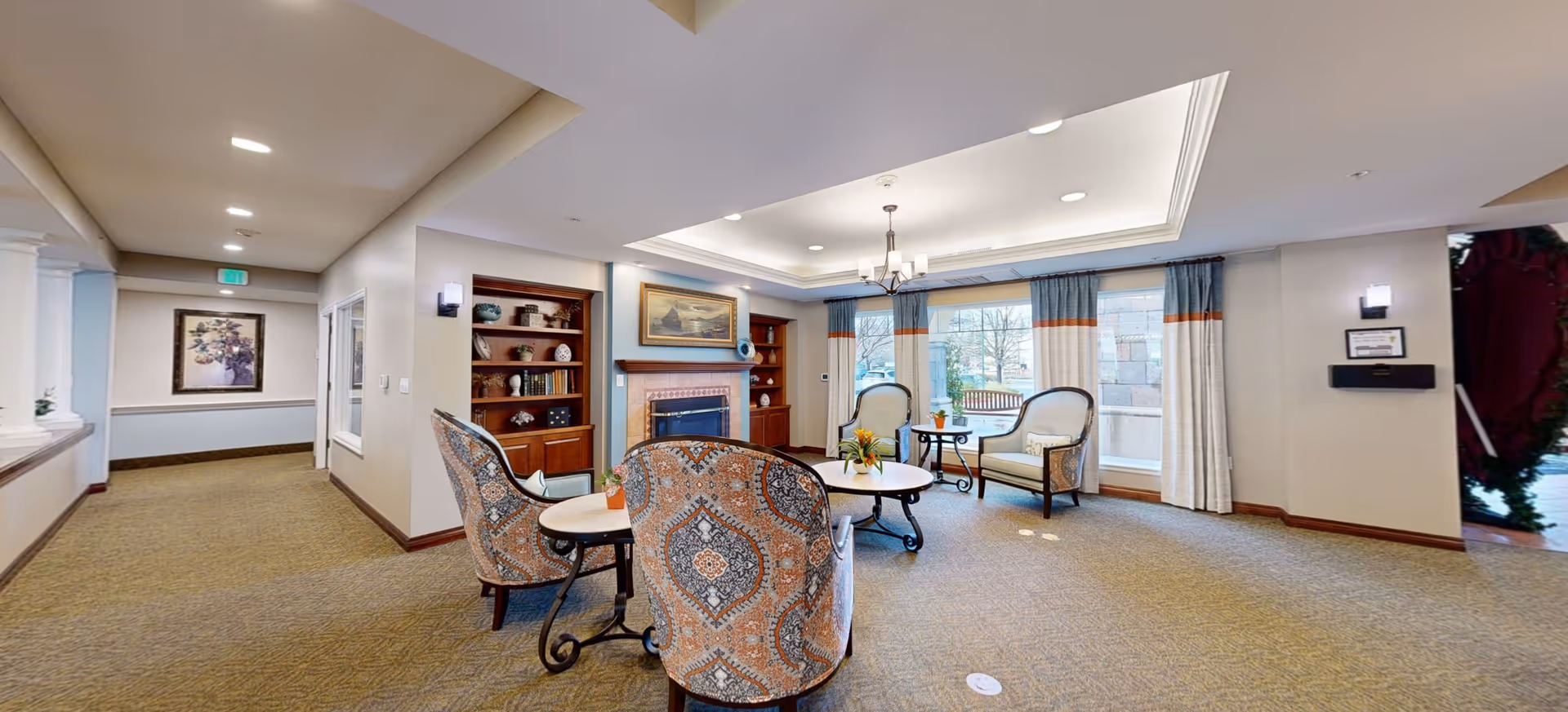 A cozy common area in a senior living facility featuring a seating arrangement with patterned armchairs and a round coffee table. The room has a fireplace with built-in wooden shelves on either side, decorated with vases and other ornaments. Large windows with curtains allow natural light to brighten the space, and a hallway extends to the left with a framed picture on the wall.