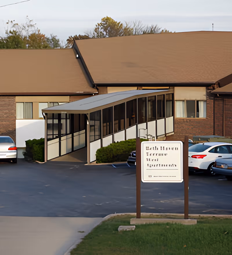 Exterior view of a single-story assisted living facility with a covered walkway leading to the entrance. Several cars are parked in the parking lot adjacent to the building. There is a sign in front of the building, and trees are visible in the background.