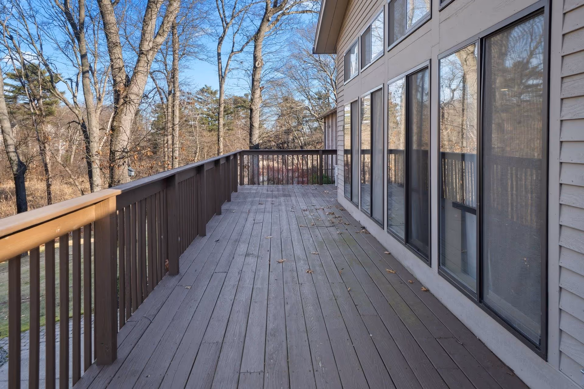 A wooden outdoor deck attached to a building with large windows reflecting the surrounding trees. The deck has a brown railing and overlooks a wooded area with leafless trees under a clear blue sky.
