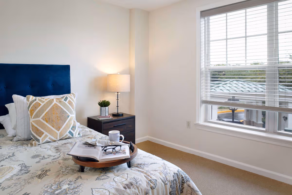 A bright bedroom with a bed featuring a blue upholstered headboard, decorative pillows, and a floral patterned bedspread. On the bed is a wooden tray holding an open book, a pair of glasses, and a white mug. Next to the bed is a dark wooden nightstand with a small potted plant, a book, and a lit table lamp. A large window with white blinds lets in natural light and shows a view of a building roof and some greenery outside.