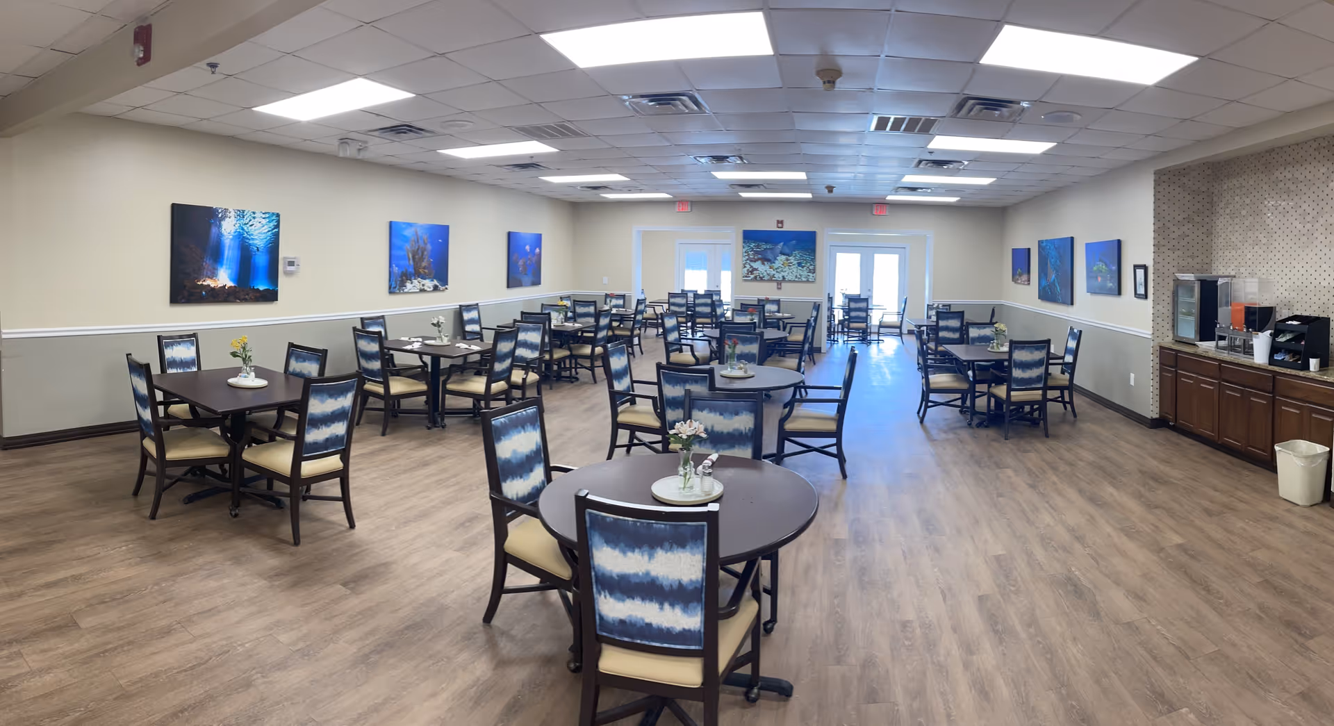 A spacious dining room with multiple round and square tables, each surrounded by chairs with blue and white patterned backs and beige seats. The room has wood flooring, beige walls with a white chair rail, and several underwater-themed paintings on the walls. On the right side, there is a counter area with a coffee machine, water dispenser, and cabinets. The ceiling has recessed lighting and ceiling tiles, and there are double doors at the far end of the room.