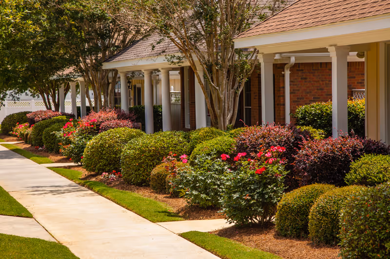 A landscaped walkway alongside a senior living facility building with neatly trimmed bushes, flowering plants, and trees. The building features brick walls, white columns, and a covered porch area.
