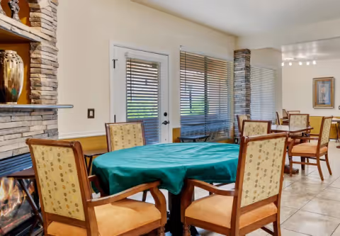 Bright communal dining area with round tables covered in green tablecloths and upholstered chairs near a fireplace and large windows.