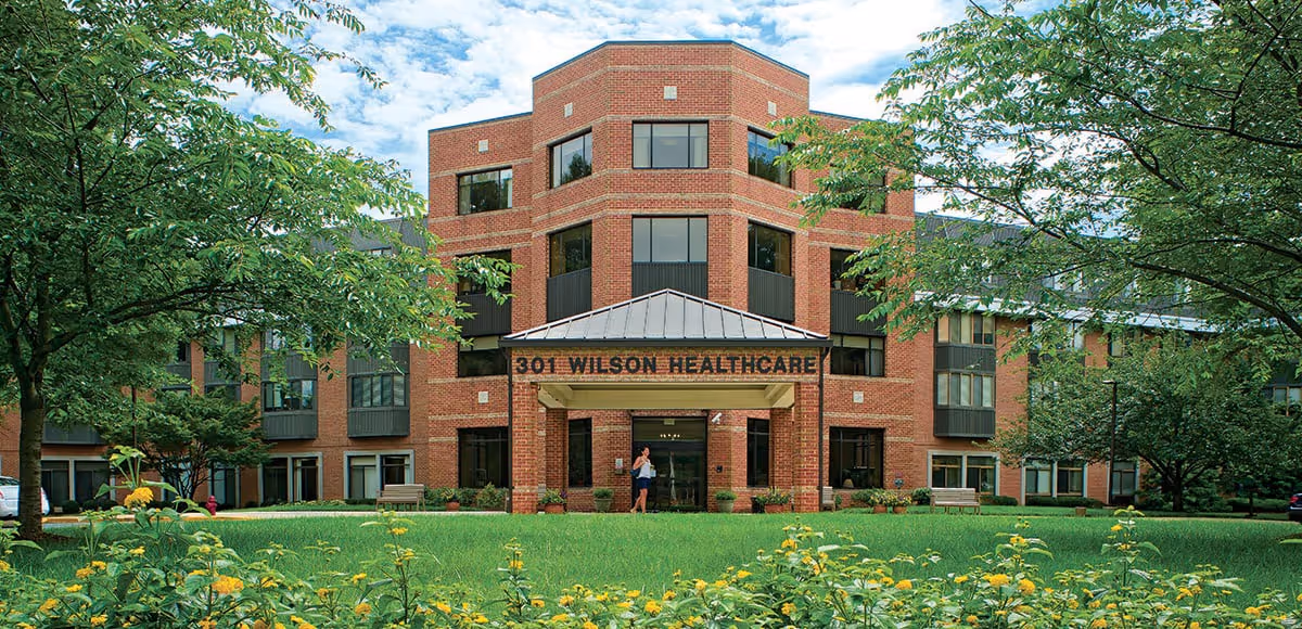 Front exterior view of a multi-story brick healthcare building with a covered entrance labeled '301 Wilson Healthcare,' surrounded by green trees, grass, and yellow flowers under a partly cloudy sky.