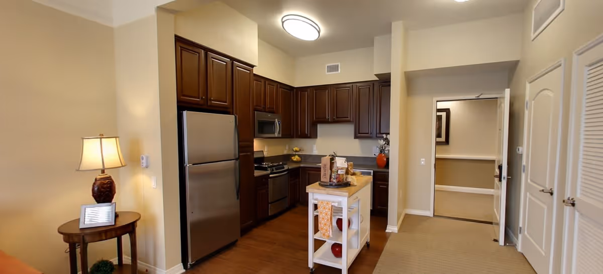 Interior view of a kitchen area in a senior living facility featuring dark wood cabinets, stainless steel refrigerator, stove, and microwave. A small white kitchen island with shelves and a towel is in the center. To the left, there is a small round table with a lamp and a framed picture. The walls are painted beige, and there is an open doorway leading to another room.
