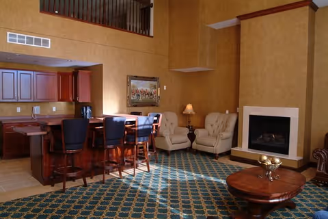 A cozy interior room with a kitchen area featuring wooden cabinets and a counter with four high chairs. Adjacent to the kitchen is a seating area with two beige armchairs, a small table with a lamp, and a fireplace. The room has a patterned carpet and warm-toned walls.